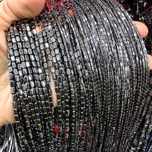 Könnte beinhalten: Eine Schnur schwarzer Hämatitperlen, jede Perle ist ein Quadrat mit abgerundeten Kanten. Die Perlen sind auf einem dünnen Draht aufgefädelt und in einem parallelen Muster angeordnet.