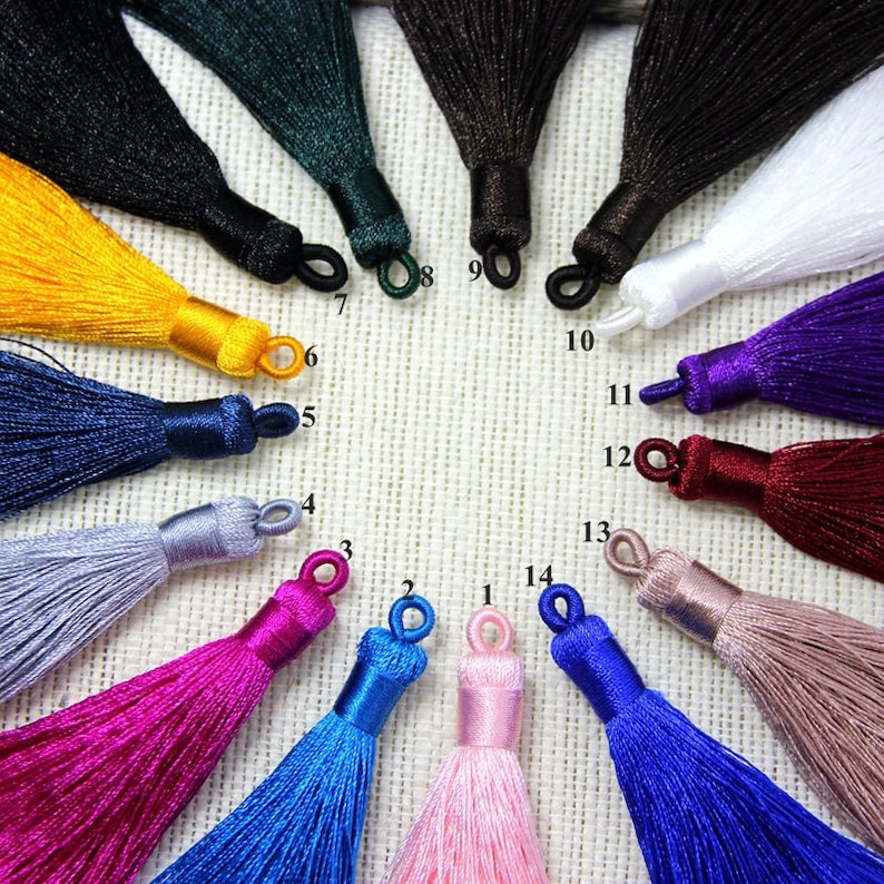 May include: Assortment of 14 different colored silk tassels with a loop at the top. The tassels are arranged in a circle on a textured fabric background.