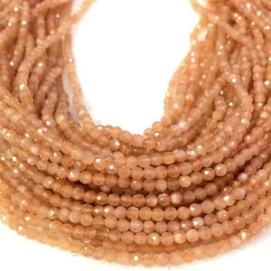 May include: Close-up of strands of small, peach-colored beads. The beads have a faceted cut, creating a sparkling effect. The beads are strung together, forming multiple strands. The background is white.