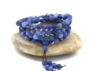 Puede incluir: Un collar mala de piedras azules con un borla. El collar está hecho de cuentas redondas y está envuelto alrededor de una piedra marrón claro.