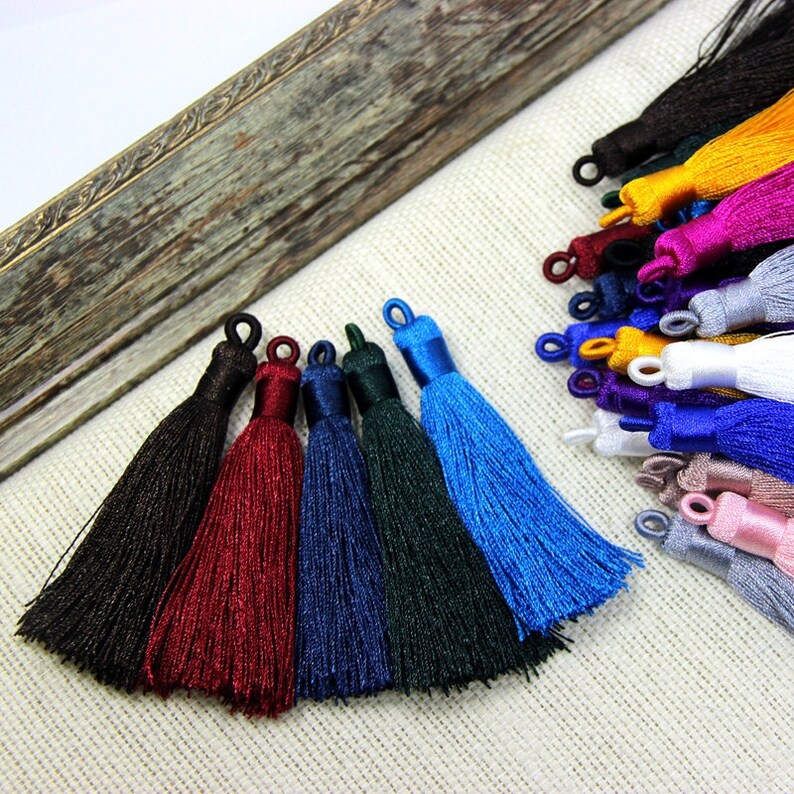 May include: Assortment of colorful silk tassels in various colors, including black, red, blue, green, and light blue. The tassels are arranged in a row on a white fabric background.
