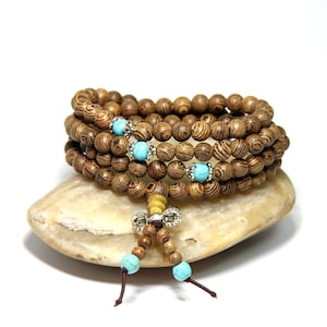 May include: A brown wooden beaded bracelet with turquoise accents and a silver charm. The bracelet is wrapped around a light brown stone.
