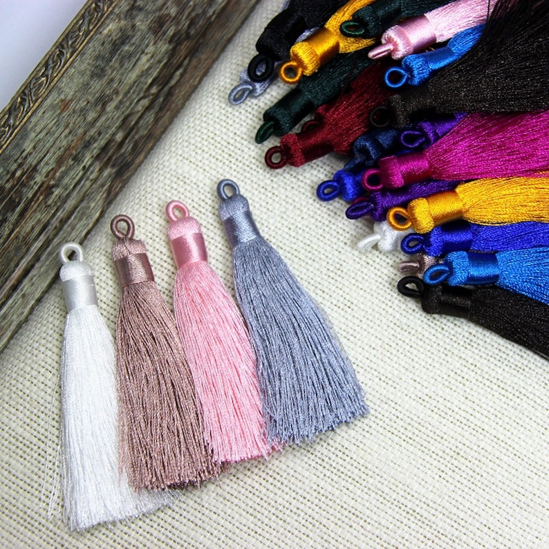 May include: Five different colored tassels, white, beige, pink, gray, and light blue, are arranged in a row on a beige fabric surface. The tassels are made of soft, silky threads and have a delicate, elegant look.