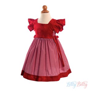 May include: Red and white gingham dress with a red velvet bodice and ruffled sleeves. The dress has a tie at the back and a red velvet trim around the bottom.