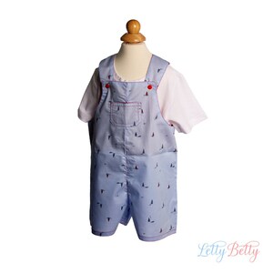 May include: A light blue overall with a white sailboat pattern and red buttons. The overall has a small pocket on the front and red stitching around the edges.