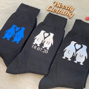 May include: Three pairs of black socks with penguin designs. The penguins are holding hearts and have initials on them. The socks also have the date 18.07.20 printed on them.