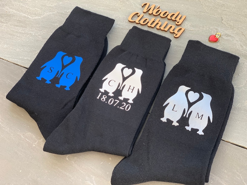 May include: Three pairs of black socks with penguin designs. The penguins are holding hearts and have initials on them. The socks also have the date 18.07.20 printed on them.