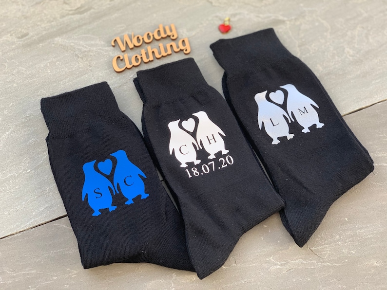 May include: Three pairs of black socks with penguin designs. The penguins are holding hearts and have initials on them. The socks also have the date 18.07.20 printed on them.