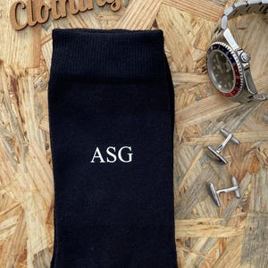 Puede incluir: Calcetines negros con las iniciales "ASG" bordadas en blanco. Los calcetines est&aacute;n sobre una superficie de madera con un reloj y gemelos.