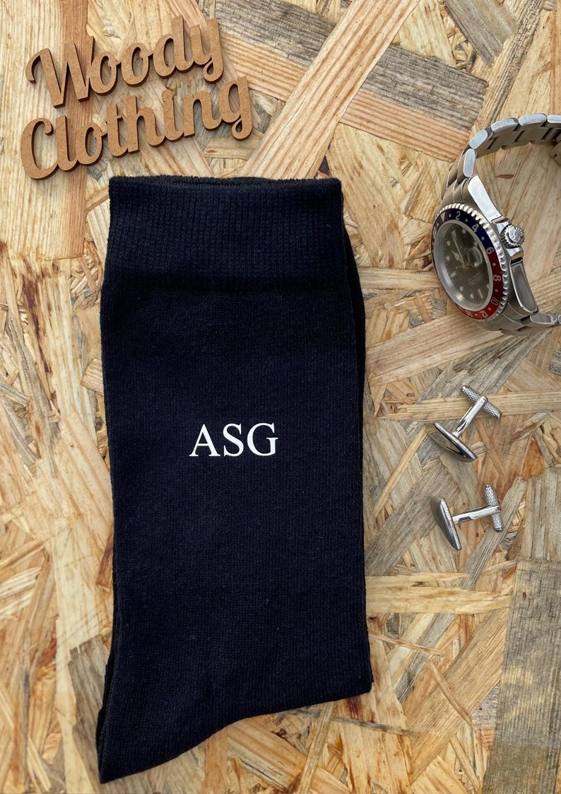 Puede incluir: Calcetines negros con las iniciales "ASG" bordadas en blanco. Los calcetines est&aacute;n sobre una superficie de madera con un reloj y gemelos.