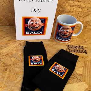May include: A Father's Day gift set featuring a white mug, black socks, and a card. The mug and socks have a cartoon character with the word "BALDI" printed on them. The card says "Happy Father's Day".