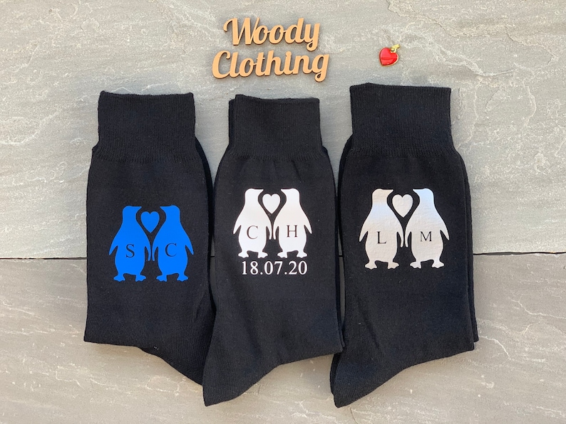 May include: Three pairs of black socks with penguin designs. The penguins are holding hearts with initials and a wedding date. The socks are a fun and unique gift for a couple.