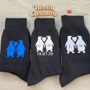 May include: Three pairs of black socks with penguin designs. The penguins are holding hearts with initials on them. The socks have the date 18.07.20 printed on them.
