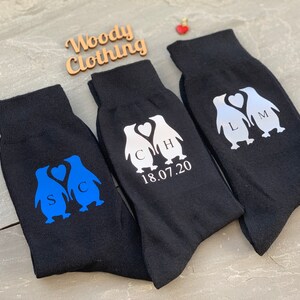 May include: Three pairs of black socks with penguin designs. The penguins are holding hearts and have initials on them. The socks also have the date 18.07.20 printed on them.