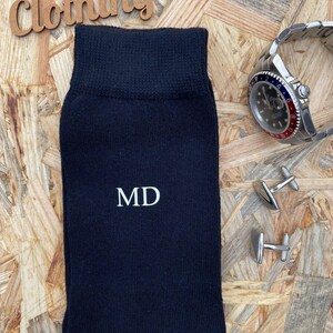 Puede incluir: Calcetines negros con las iniciales "MD" bordadas en blanco en la parte delantera. Los calcetines est&aacute;n sobre una superficie de madera con un reloj y gemelos.