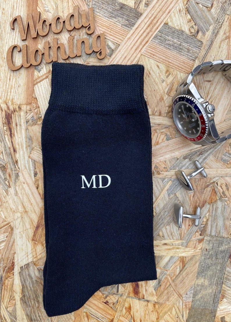 Puede incluir: Calcetines negros con las iniciales "MD" bordadas en blanco en la parte delantera. Los calcetines est&aacute;n sobre una superficie de madera con un reloj y gemelos.
