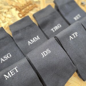 Puede incluir: Un juego de ocho calcetines de color gris oscuro con iniciales bordadas en blanco. Las iniciales son: ASG, MET, AMM, JDS, ATP, TJRG, RET y RAS.