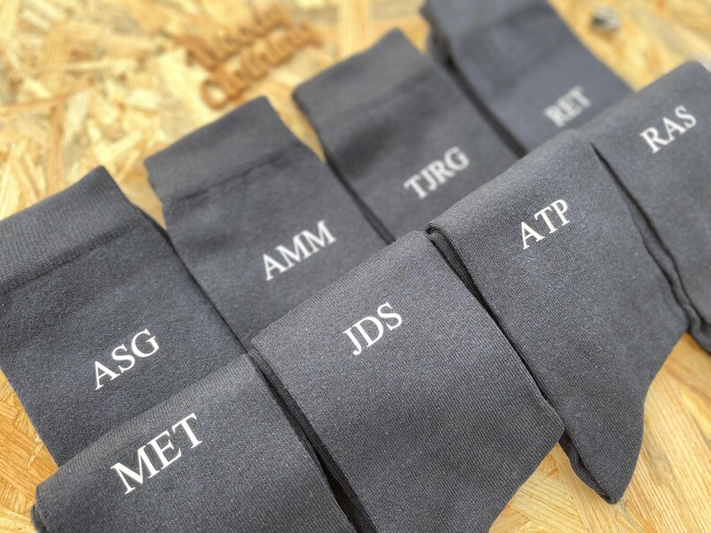 Puede incluir: Un juego de ocho calcetines de color gris oscuro con iniciales bordadas en blanco. Las iniciales son: ASG, MET, AMM, JDS, ATP, TJRG, RET y RAS.
