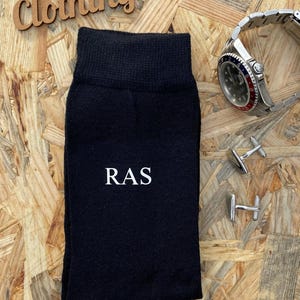 Puede incluir: Calcetines negros con las iniciales "RAS" bordadas en blanco en la parte delantera. Los calcetines est&aacute;n sobre un fondo de madera con gemelos y un reloj.