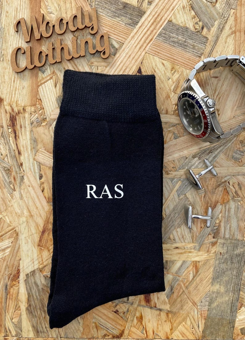 Puede incluir: Calcetines negros con las iniciales "RAS" bordadas en blanco en la parte delantera. Los calcetines est&aacute;n sobre un fondo de madera con gemelos y un reloj.