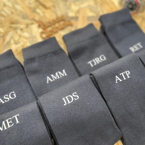 Puede incluir: Un juego de diez calcetines de color gris oscuro con iniciales bordadas en blanco. Las iniciales son: ASG, MET, AMM, JDS, TJRG, ATP, RET, RAS.