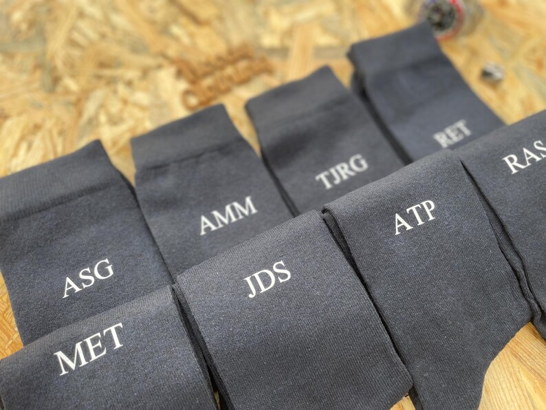 Puede incluir: Un juego de diez calcetines de color gris oscuro con iniciales bordadas en blanco. Las iniciales son: ASG, MET, AMM, JDS, TJRG, ATP, RET, RAS.