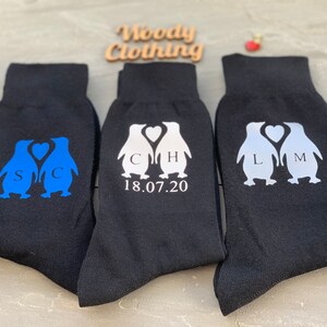 May include: Three pairs of black socks with penguin designs. The penguins are holding hearts and have initials on their chests. One pair has blue penguins, one pair has white penguins, and one pair has silver penguins. The socks also have the date 18.07.20 printed on them.