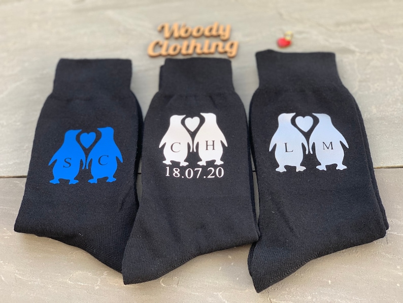 May include: Three pairs of black socks with penguin designs. The penguins are holding hearts and have initials on their chests. One pair has blue penguins, one pair has white penguins, and one pair has silver penguins. The socks also have the date 18.07.20 printed on them.