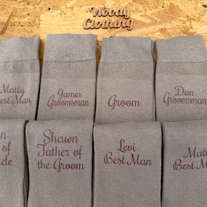 May include: Eight pairs of grey socks, each embroidered with a different title in burgundy thread. Titles include "Matty Best Man", "James Groomsman", "Groom", "Dan Groomsman", "Brian Father of the Bride", "Shawn Father of the Groom", "Levi Best Man", and "Matt Best Man".