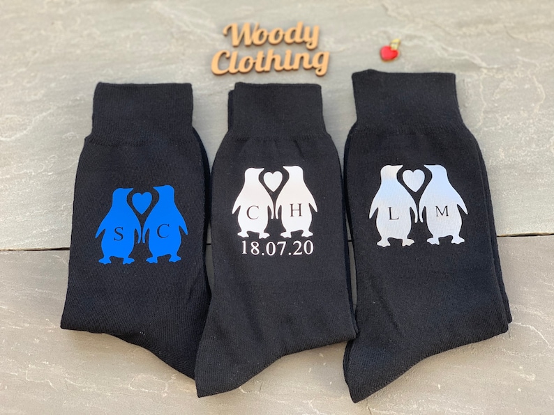 May include: Three pairs of black socks with penguin designs. The penguins are holding hearts with initials on them. The socks have the date 18.07.20 printed on them.