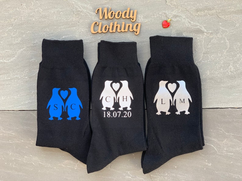 May include: Three pairs of black socks with penguin designs. The penguins are holding hearts with initials and a wedding date. The socks are a fun and unique gift for a couple.