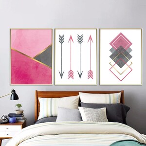 May include: Set of three abstract wall art prints featuring geometric shapes and arrows in pink, gray, and gold. The prints are framed in a gold frame.