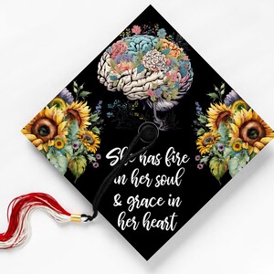 May include: Black graduation cap with a floral brain design and sunflowers. The cap features the quote, "She has fire in her soul & grace in her heart" in white script. A red and white tassel hangs from the cap.