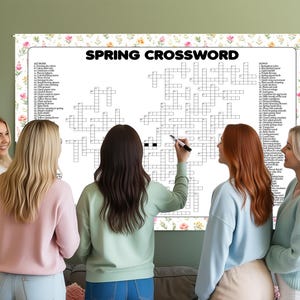 May include: A large white "Spring Crossword" puzzle with floral border is displayed. Several people are gathered around the puzzle, with one person holding a pen. The crossword has clues and spaces for answers. The background is a light green wall.