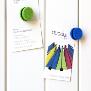 May include: Two business cards with the text "quadu unikate handgearbeitet" and a logo of colorful tents. One card has a green felt magnet and the other has a blue felt magnet.