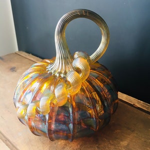 May include: A glass pumpkin with a swirl design in amber and iridescent colors. The pumpkin has a twisted handle.