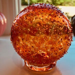 May include: A decorative, spherical glass object with an orange and amber color scheme. The surface is textured with small, raised bumps, and the base is clear. The glass sphere sits on a light-colored surface, with a blurred background.