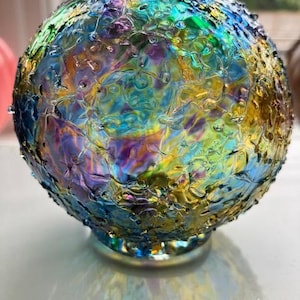 May include: A decorative glass orb with a textured surface. The orb features a vibrant display of colors, including blue, green, yellow, and purple. It sits on a small, clear base, and is suitable for home decor.