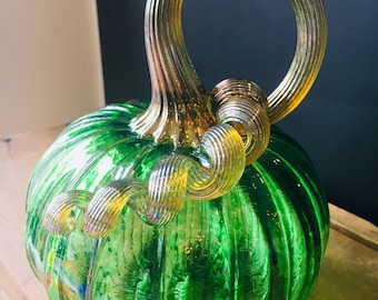 May include: A green glass pumpkin with a gold swirl stem. The pumpkin has a ribbed texture and is a decorative piece.