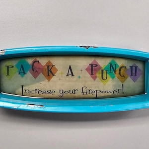 May include: A vintage-style, turquoise-colored sign with the words "PACK A PUNCH" and "Increase your firepower!" in a retro font. The sign features colorful diamond shapes and a distressed finish.