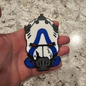 May include: A white, blue, and black mask-shaped accessory. The design features a white base with a blue central element, black accents, and red details. The accessory has a detailed, mechanical appearance.