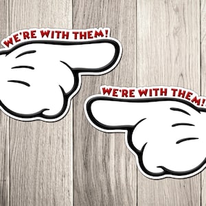 May include: Two white cartoon hands with black outlines and the text "WE'RE WITH THEM!" in red. The hands are pointing to the right. The background is a light brown wood grain.