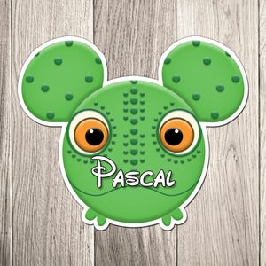May include: A green sticker featuring a cartoon frog with large orange eyes and the name "Pascal" in white script. The frog has a round head with two Mickey Mouse-shaped ears. The background is a wooden surface.