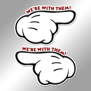 May include: Two white cartoon-style gloved hands with black outlines, each pointing in opposite directions. Above each hand, the text WE'RE WITH THEM! is printed in bold red capital letters. The design features a thick white border.