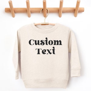 May include: A beige sweatshirt with long sleeves and a crew neck. The sweatshirt has black text that says "Custom Text" in a retro font.