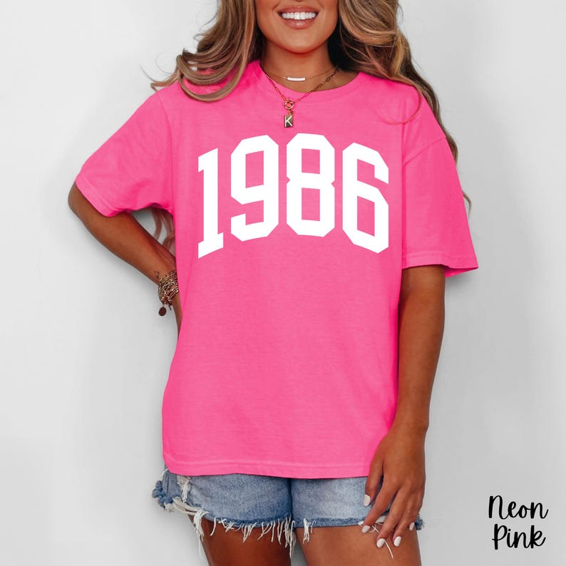 May include: A neon pink t-shirt with the year "1986" printed in large, white, block letters across the front. The shirt is a vibrant, solid color and has short sleeves. The words "Neon Pink" are in the bottom right corner.