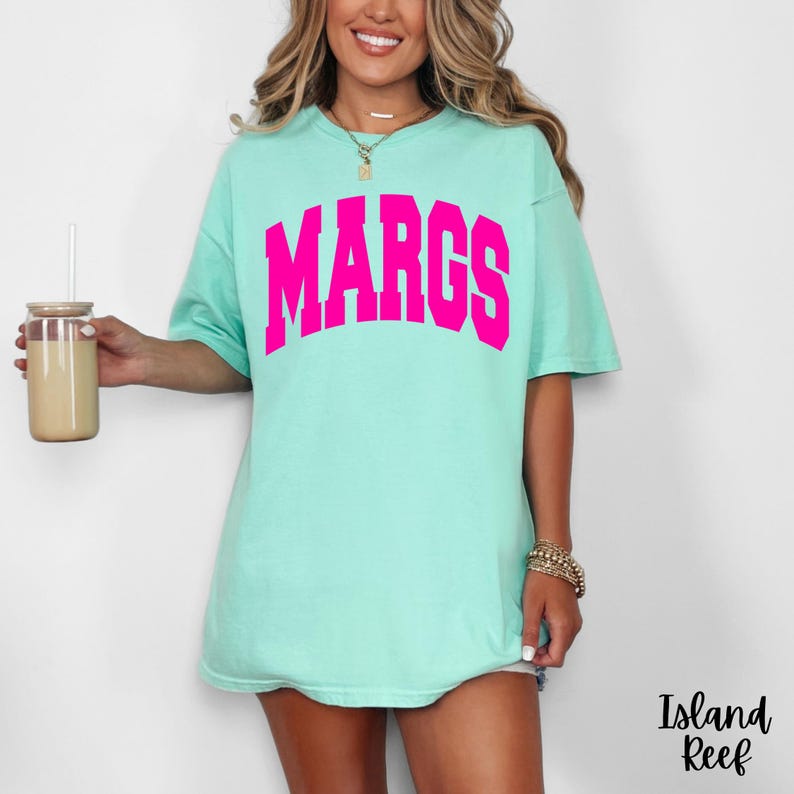 May include: A light turquoise t-shirt with the word "MARGS" in bright pink block letters. The shirt is a relaxed fit. The image also shows a clear glass with a straw and a drink inside, and the text "Island Reef" in the bottom right corner.