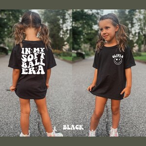 May include: Black t-shirt with white text that says "In My Soft Ball Era" on the back and a white smiley face with the text "Olivia" on the front.