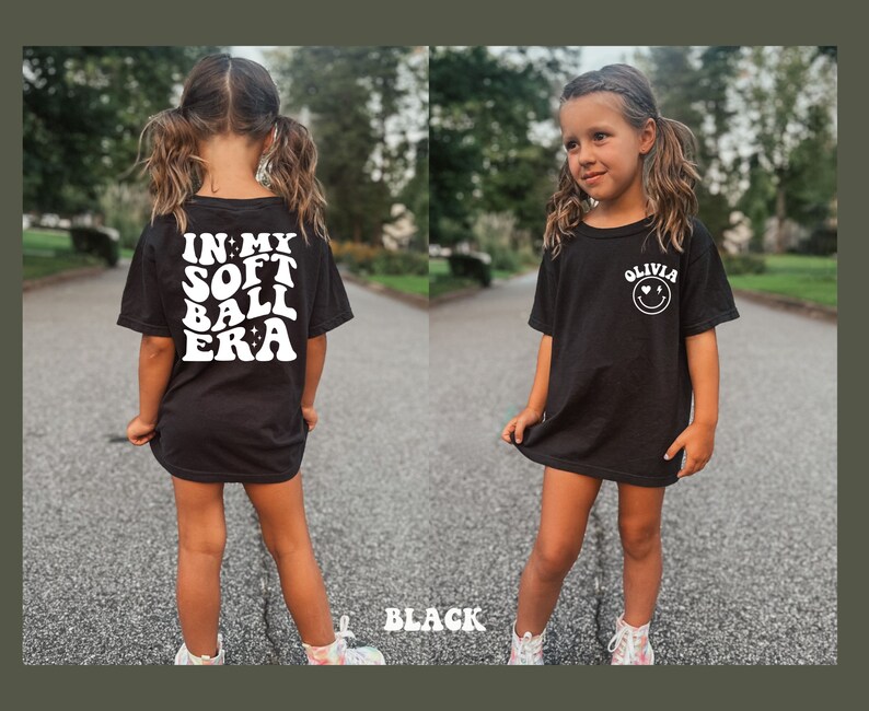 May include: Black t-shirt with white text that says "In My Soft Ball Era" on the back and a white smiley face with the text "Olivia" on the front.