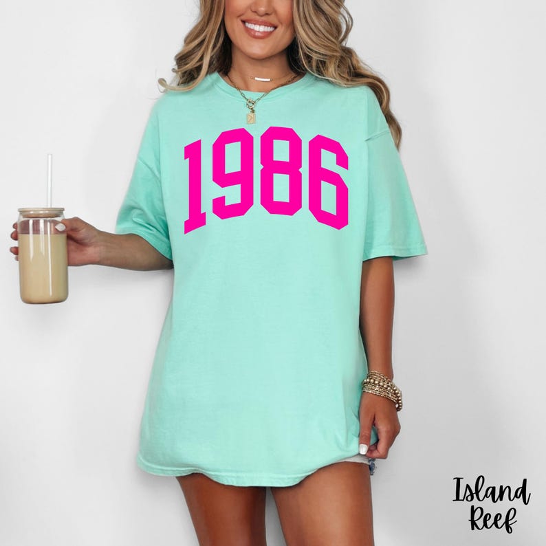 May include: A light turquoise t-shirt with the year "1986" printed in bright pink. The shirt has short sleeves and a relaxed fit. The text "Island Reef" is printed in black script in the lower right corner.
